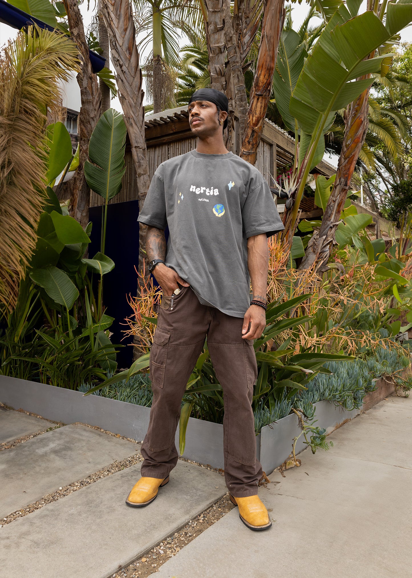 Man standing outdoors among plants wearing a gray t-shirt and brown pants.