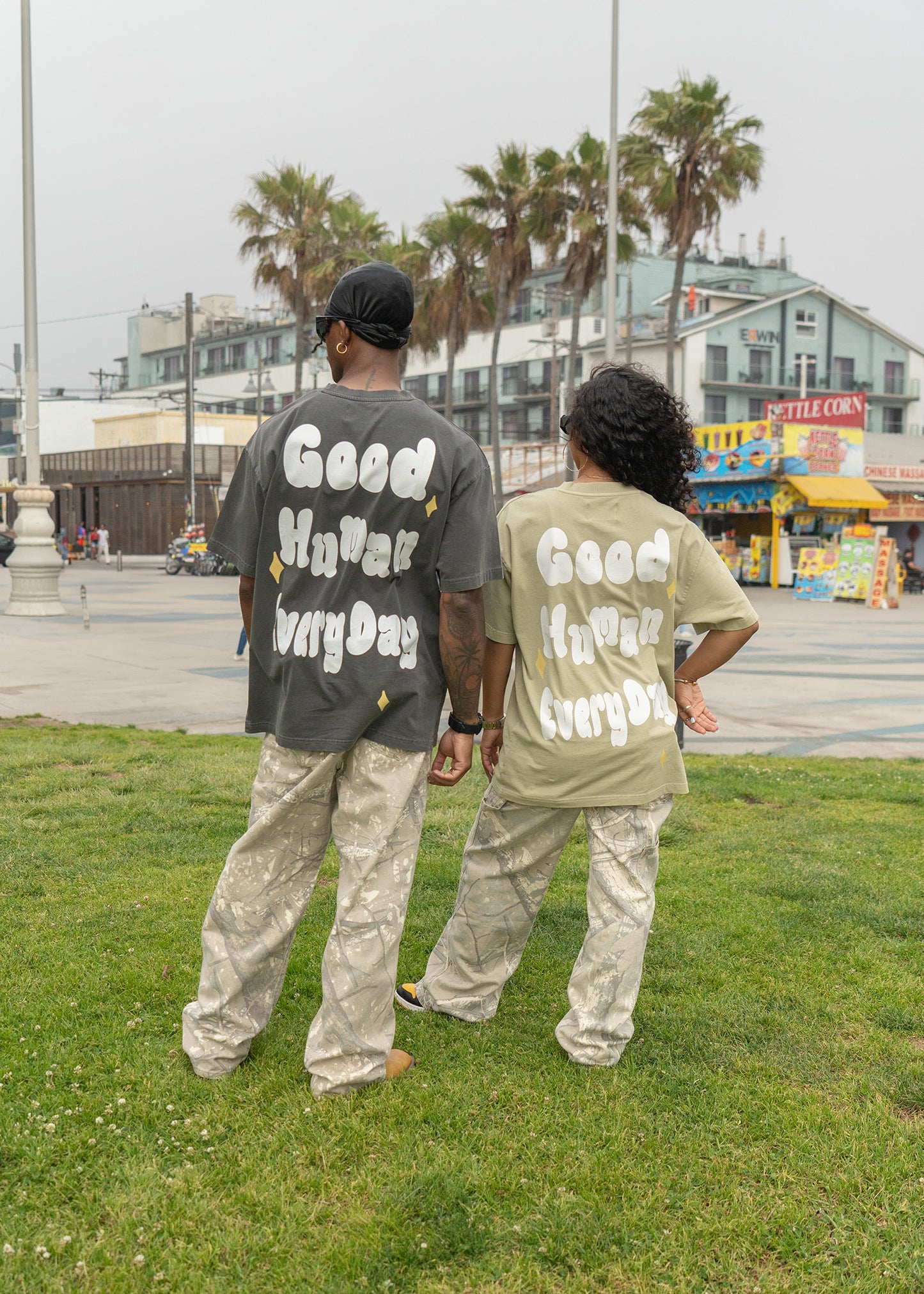 Two people walking on grass with text on their shirts, palm trees and buildings in the background.