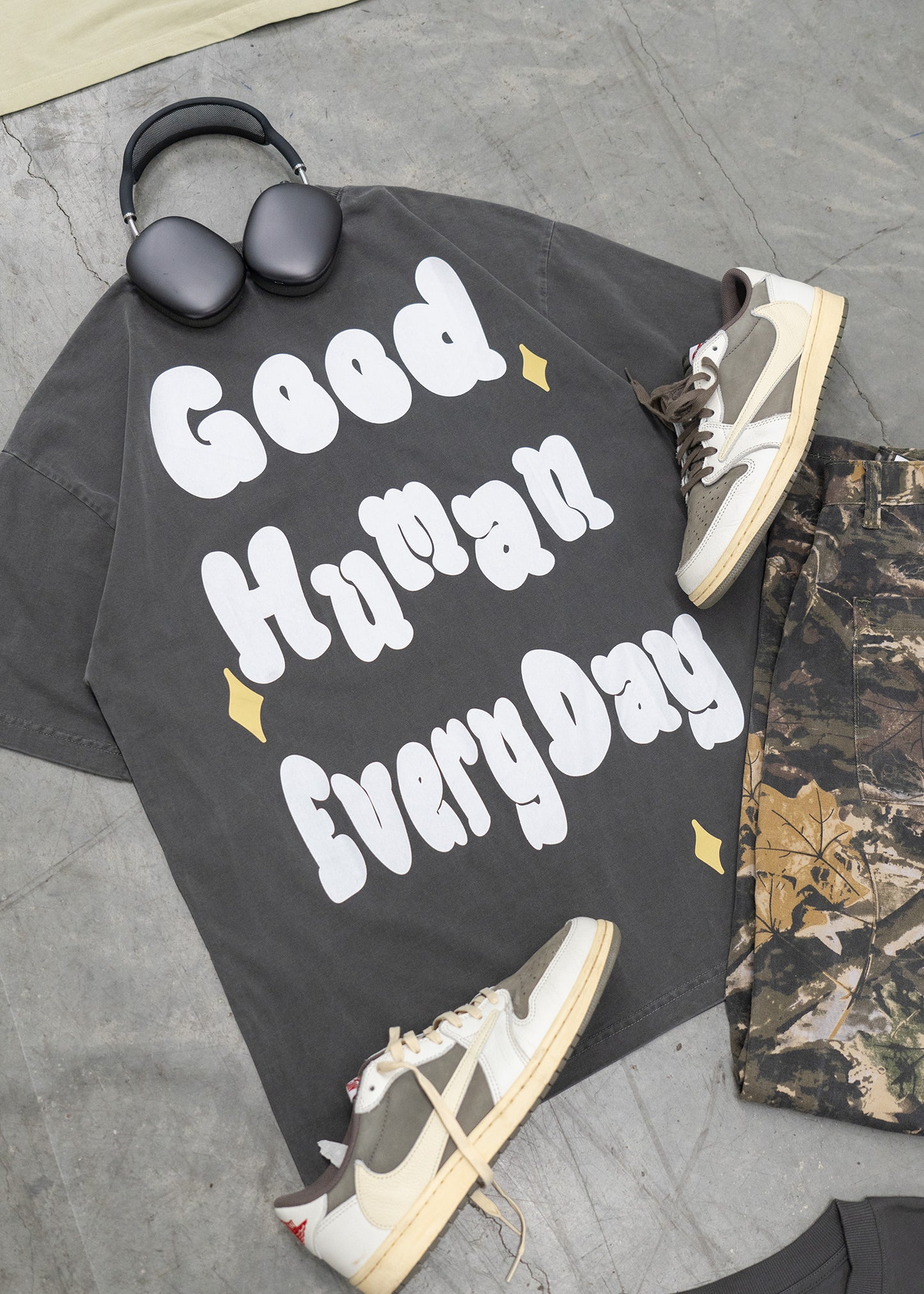 Black t-shirt with 'Good Human Every Day' text, sneakers, and a bag on a gray pavement.
