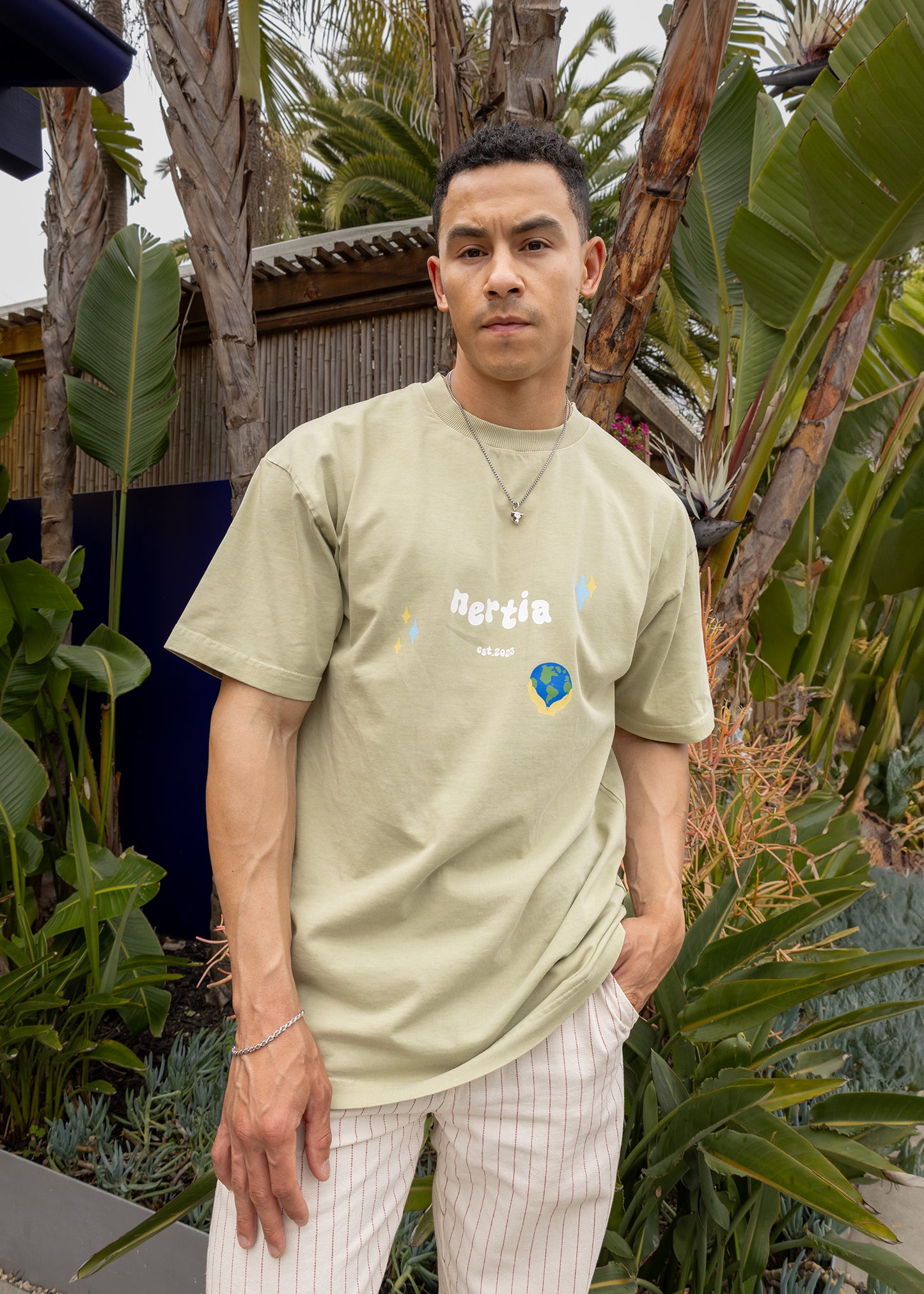 Man wearing a light green t-shirt with text and graphics, standing outdoors among plants.