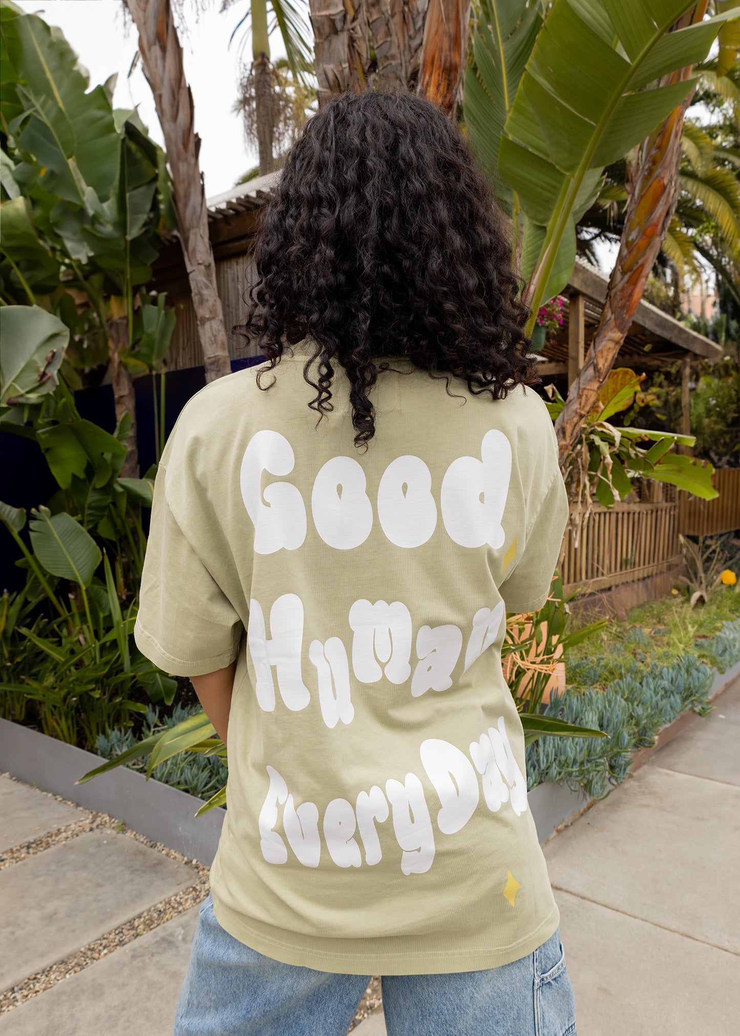 Person wearing a light green t-shirt with text on the back, standing outdoors with plants in the background