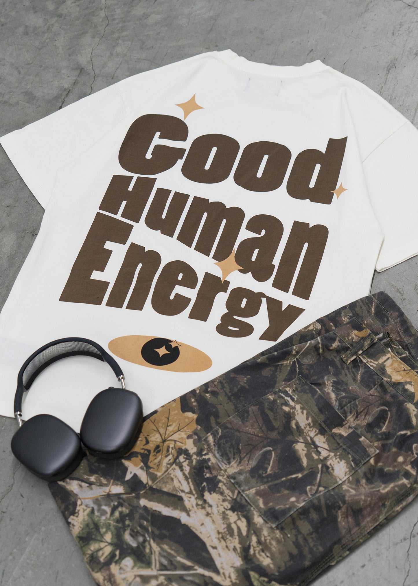 White t-shirt with 'Good Human Energy' text, black headphones, and camouflage bag on a gray surface