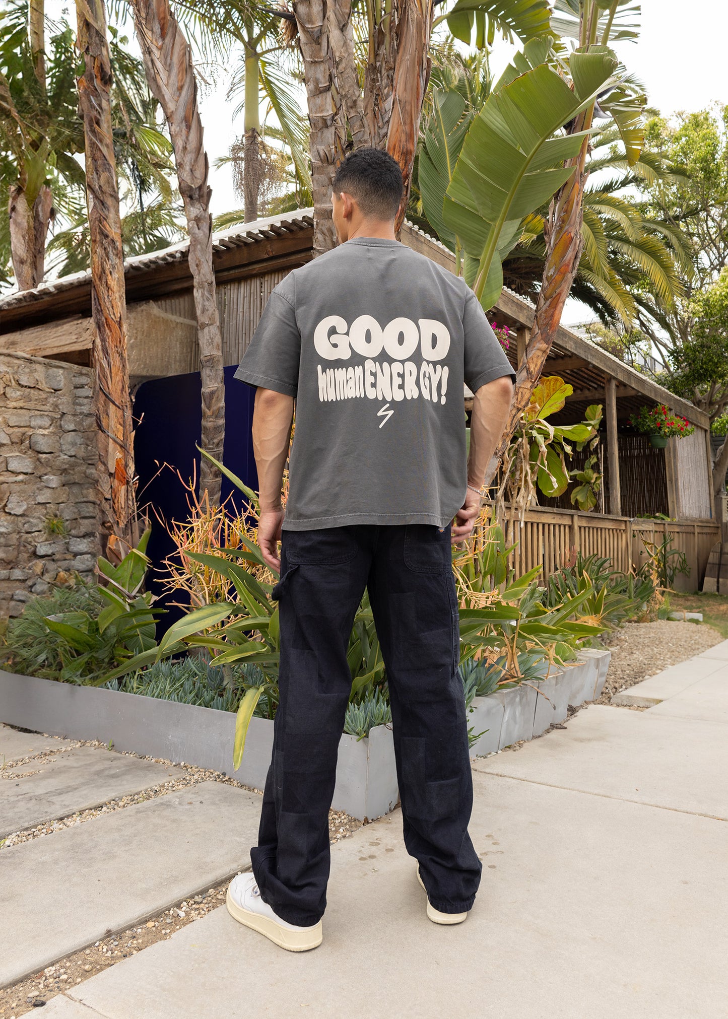 Person wearing a gray t-shirt with 'GOOD human ENERGY' text, standing outdoors with greenery and a building in the background.