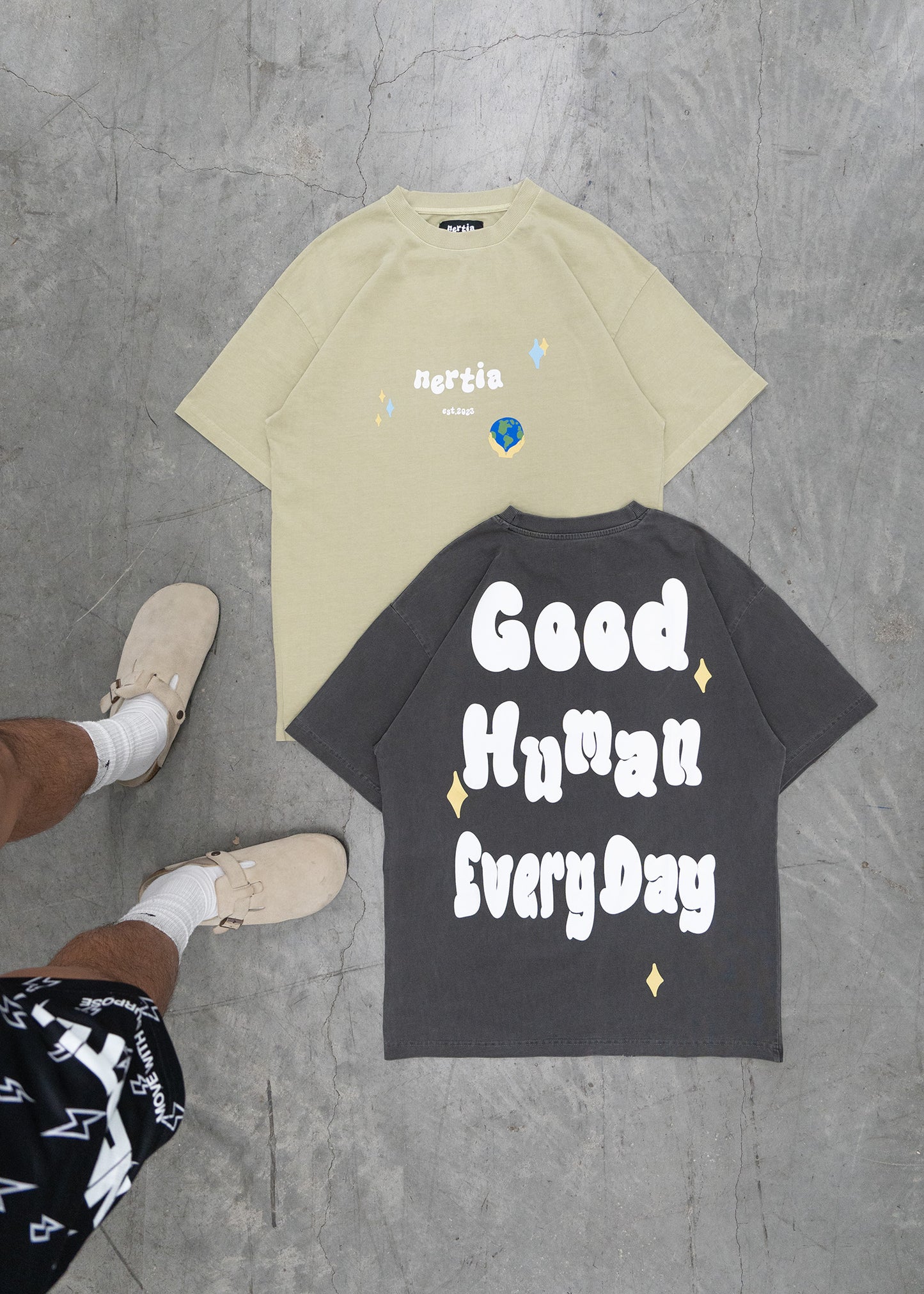 Two t-shirts on a concrete floor, one beige with text and one black with 'Good Human Every Day' text.