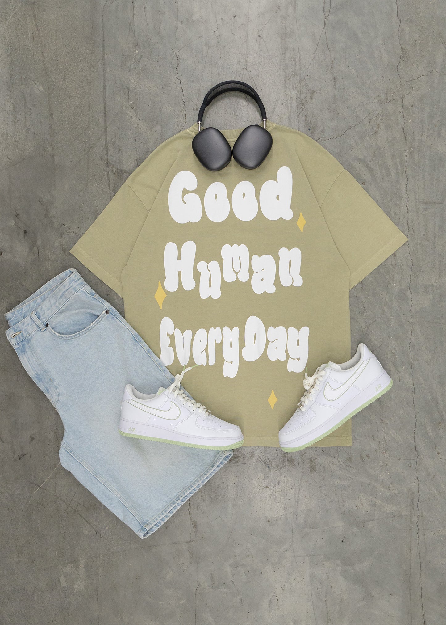 T-shirt with 'Good Human Every Day' text, jeans, sneakers, and sunglasses on a concrete floor.