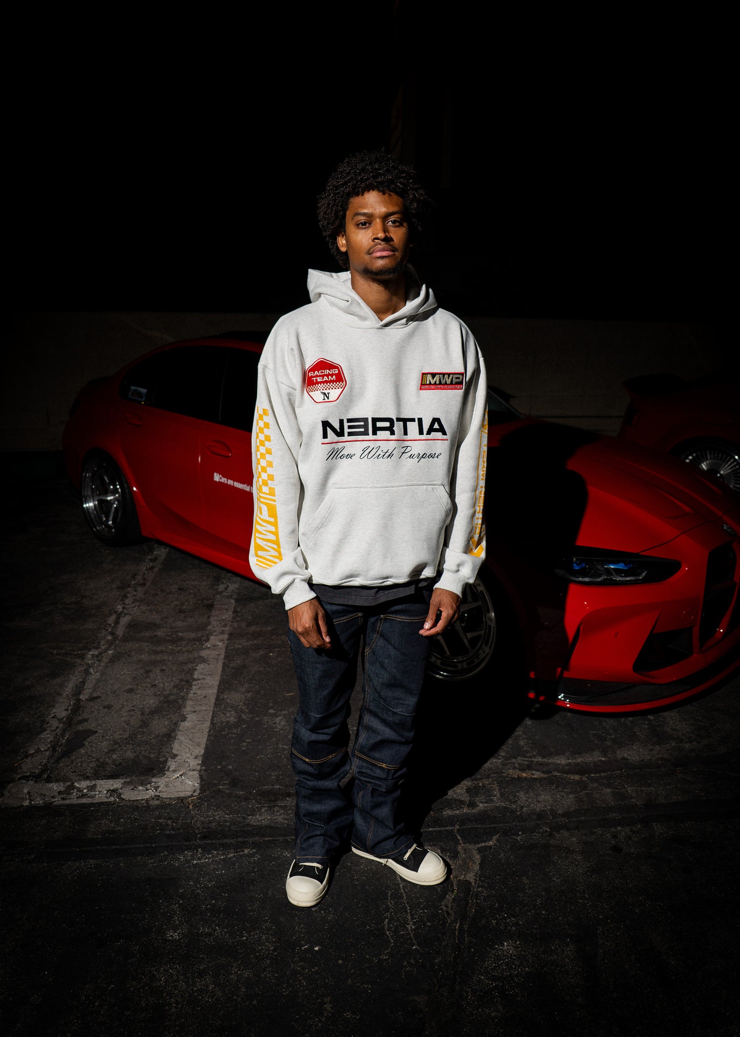 Person wearing a white hoodie with branding in front of a red car