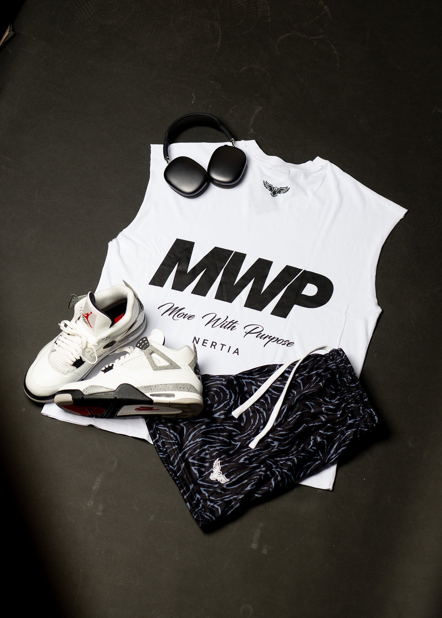 White tank top with 'MWP' text, sneakers, sunglasses, and shorts on a dark surface