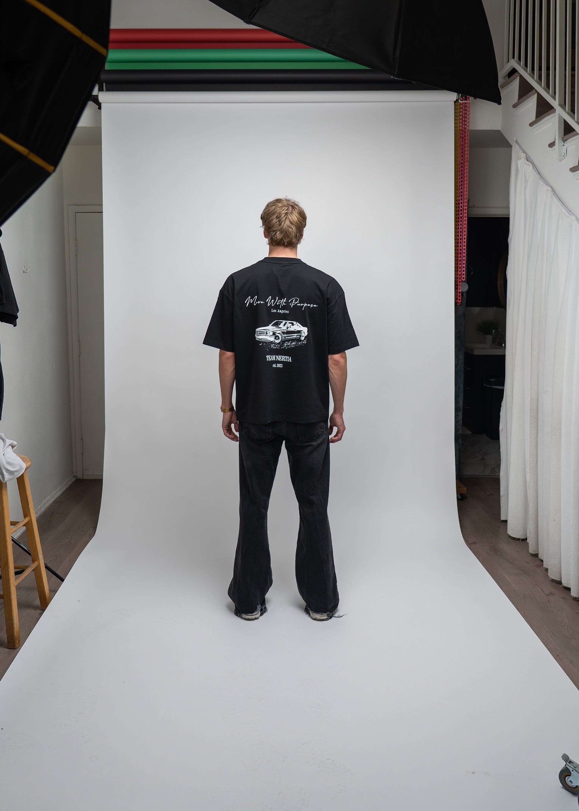 Person wearing a black t-shirt with a logo on the back, standing in front of a white backdrop in a studio setting.