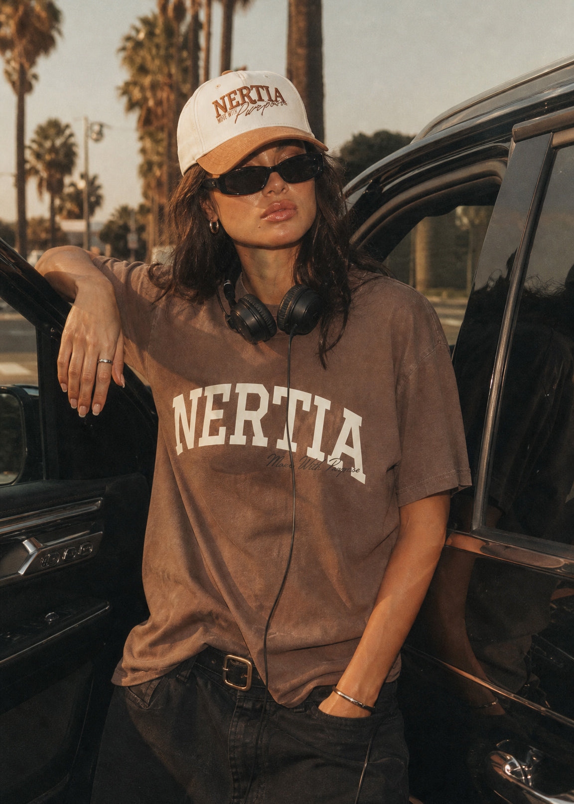 Person wearing a 'NERTIA' t-shirt and cap, leaning against a car with palm trees in the background.