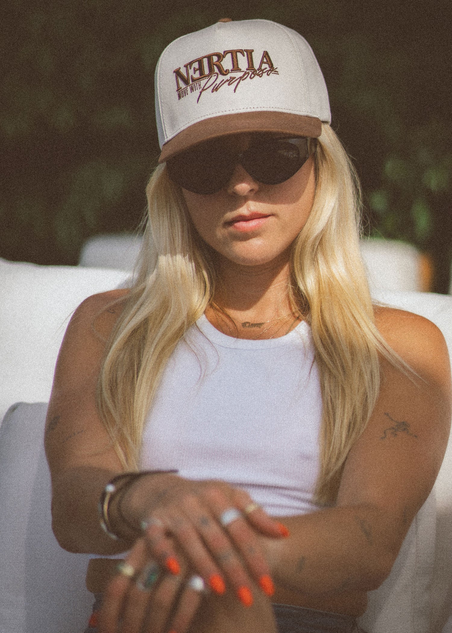 Woman wearing a white cap with 'Neruda' branding, sunglasses, and a white tank top outdoors.