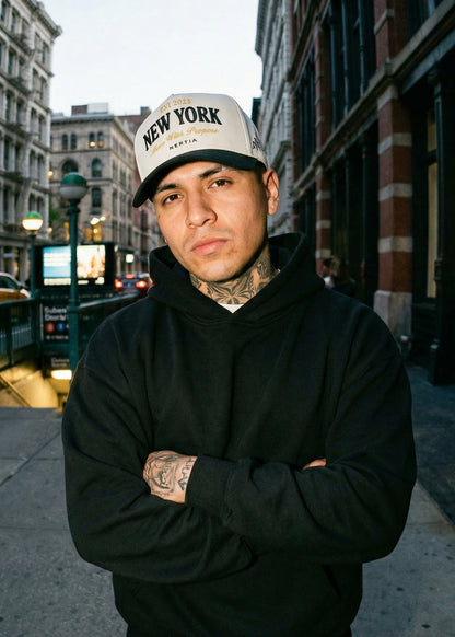 Man wearing a black hoodie and 'New York' cap standing on a city street.