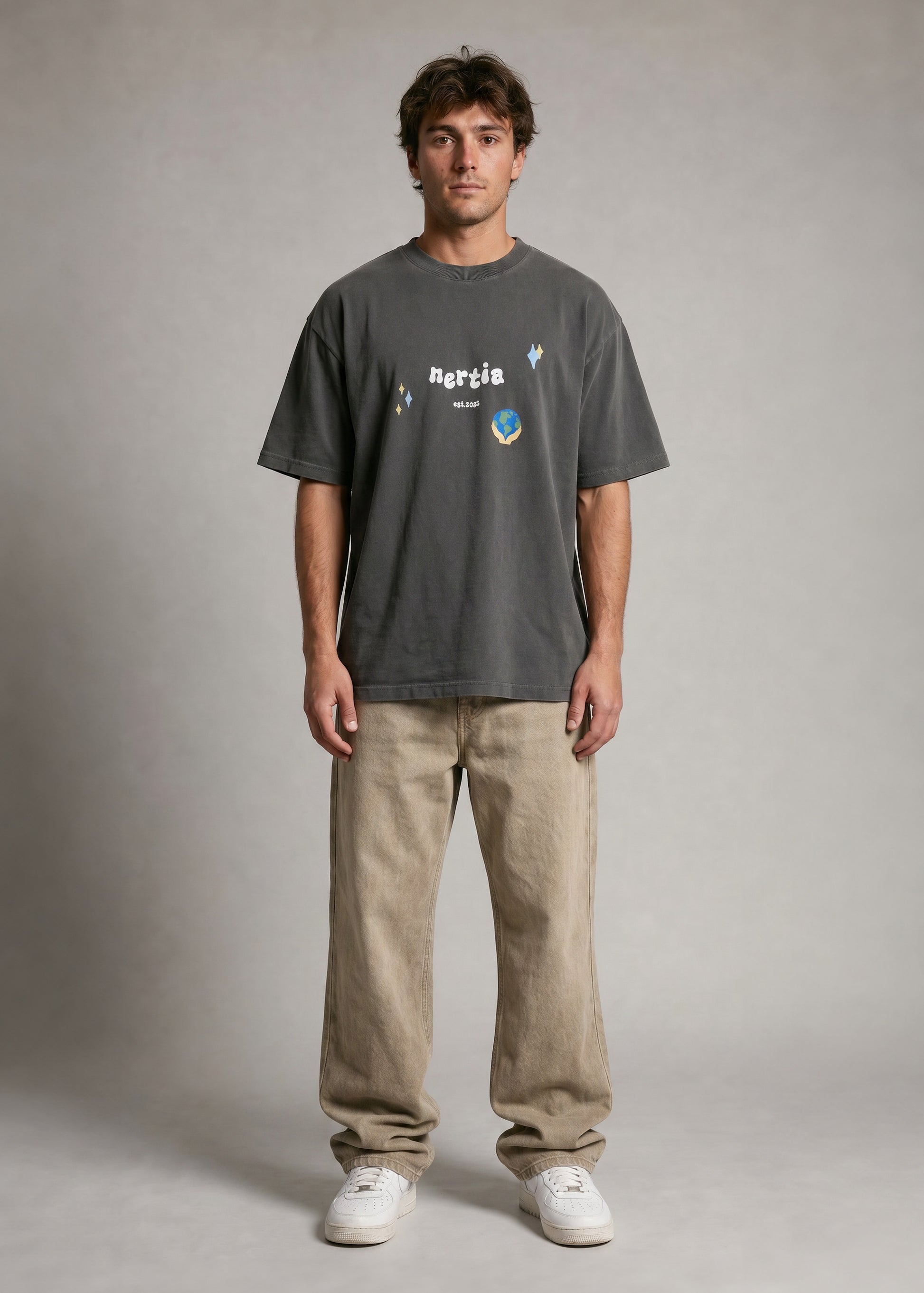 Man wearing a dark gray t-shirt with a logo and text, beige pants, and white shoes on a plain background