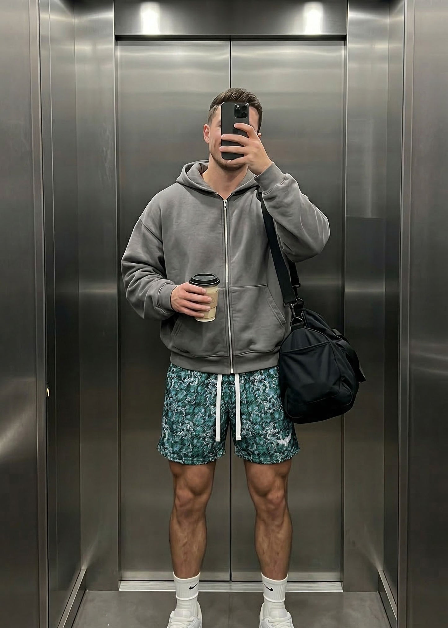 Person taking a mirror selfie in an elevator wearing a gray hoodie, patterned shorts, and white sneakers.