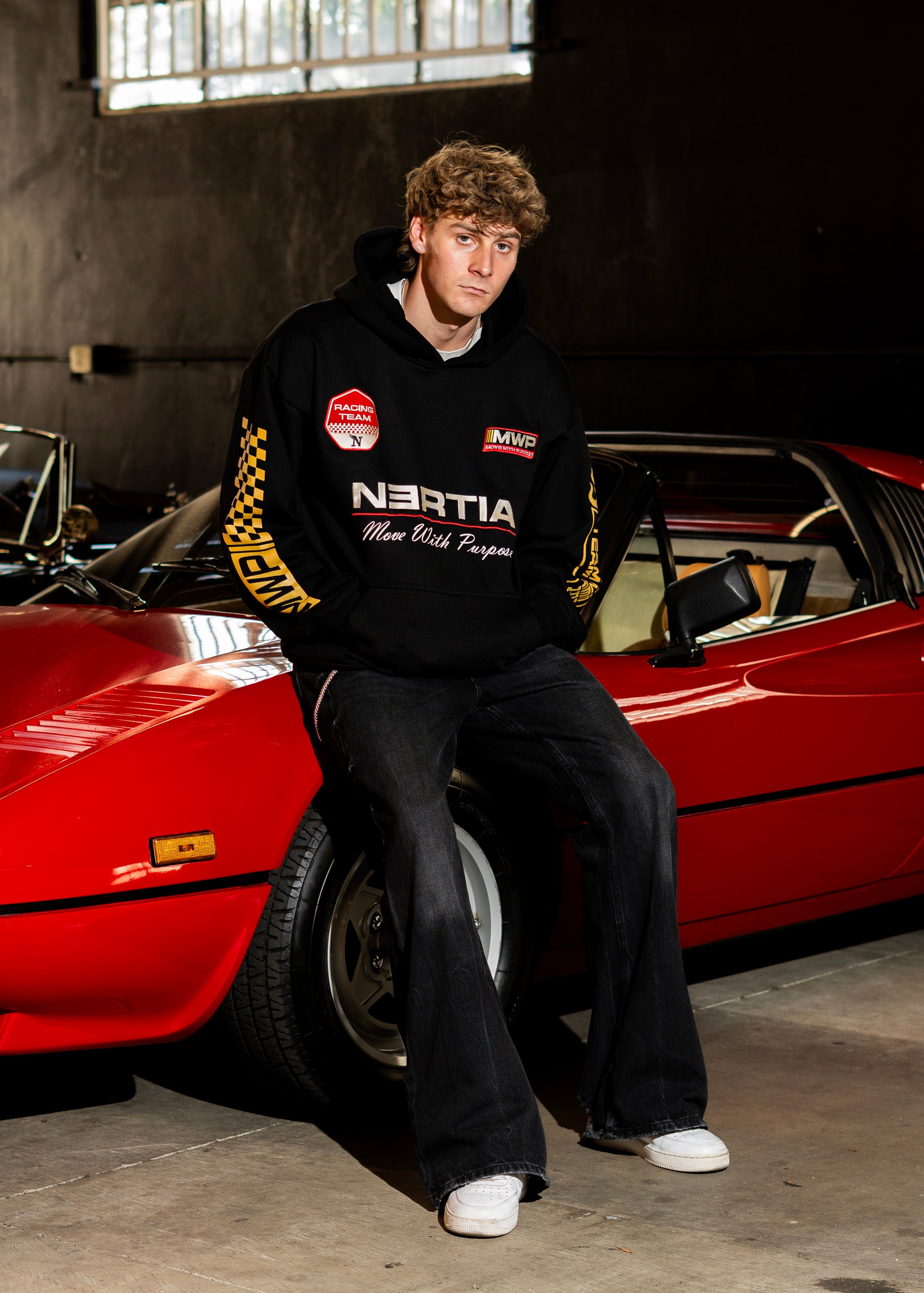 Person wearing a black hoodie with logos sitting on a red car in a garage.