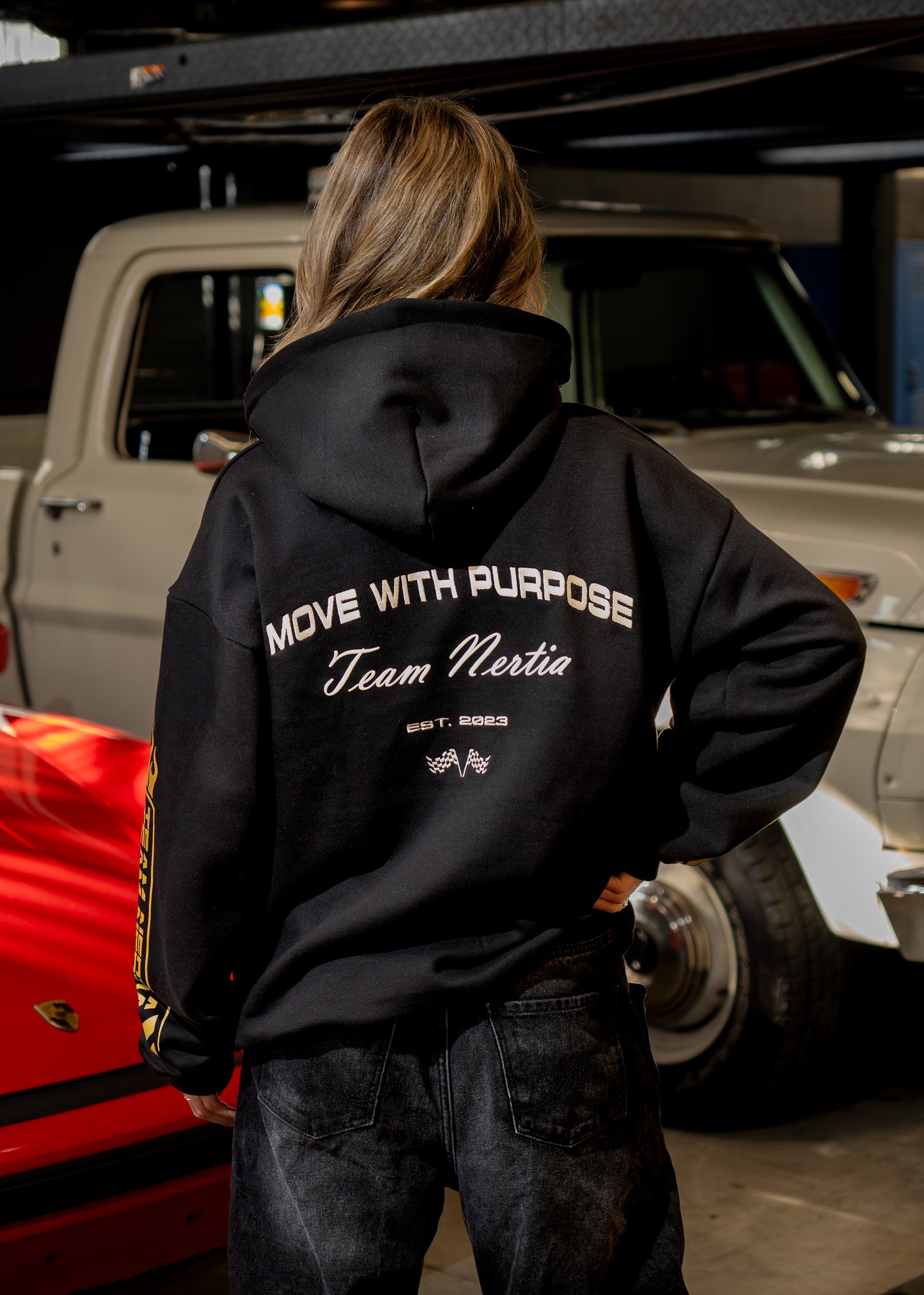 Person wearing a black hoodie with text, standing in front of a red car and beige truck.