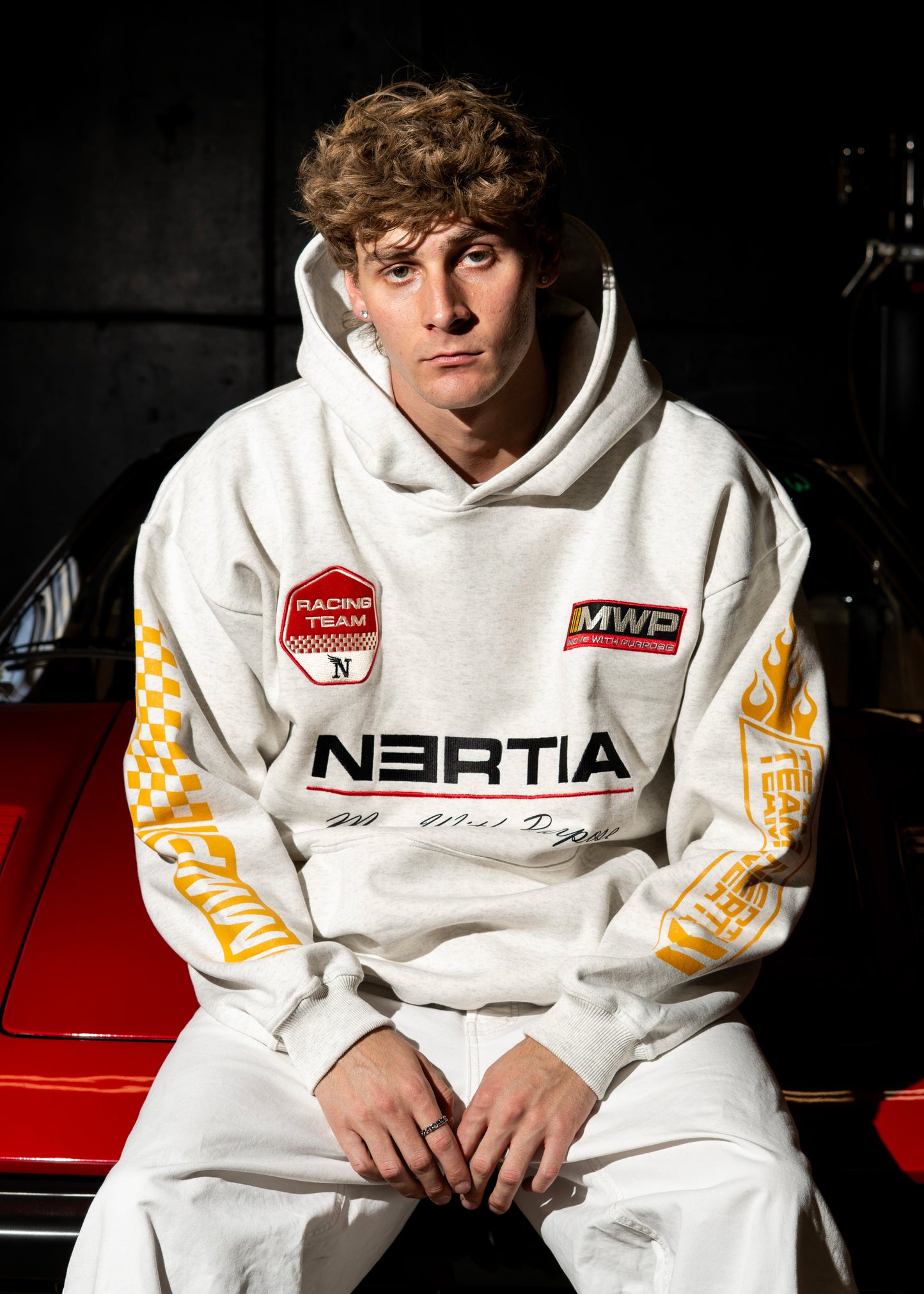 Person wearing a white hoodie with branding sitting in front of a red car.