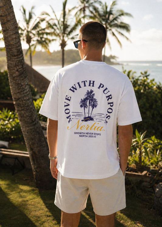 Man wearing a white t-shirt with a palm tree design and text, standing outdoors with palm trees and water in the background.