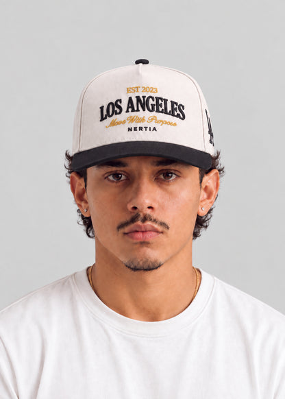 A man wearing a white Los Angeles baseball cap with black and gold embroidery, paired with a white t-shirt, photographed against a neutral gray background.