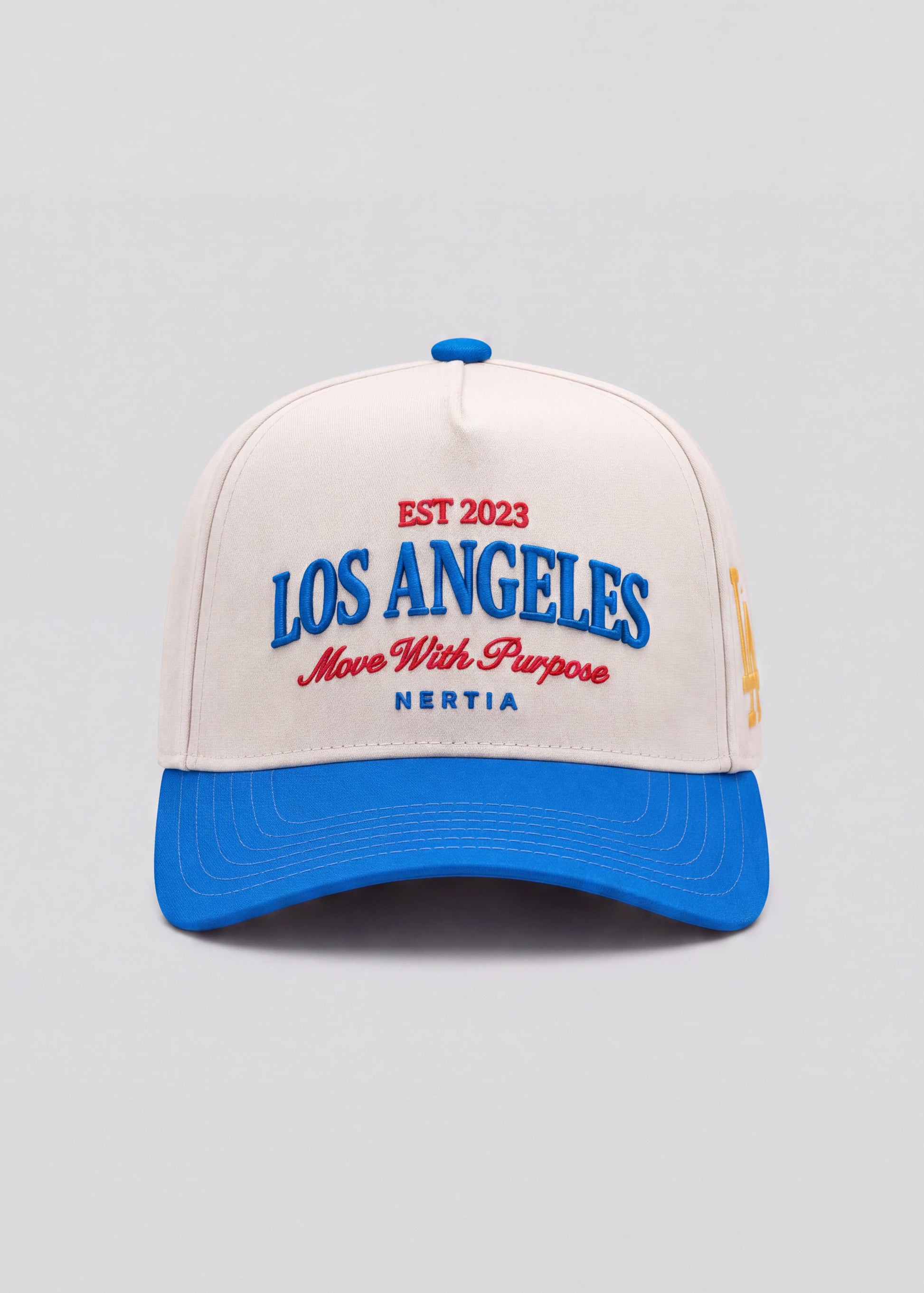 A structured baseball cap with an off-white crown and a bright blue curved brim. The front features embroidered text reading 'EST 2023 LOS ANGELES Move With Purpose NERTIA' in blue and red.