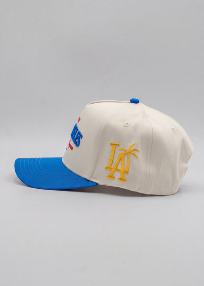 An off-white baseball cap with a bright blue visor and blue button, featuring a yellow embroidered 'LA' with a palm tree on the front and a blue and red embroidered logo on the side.