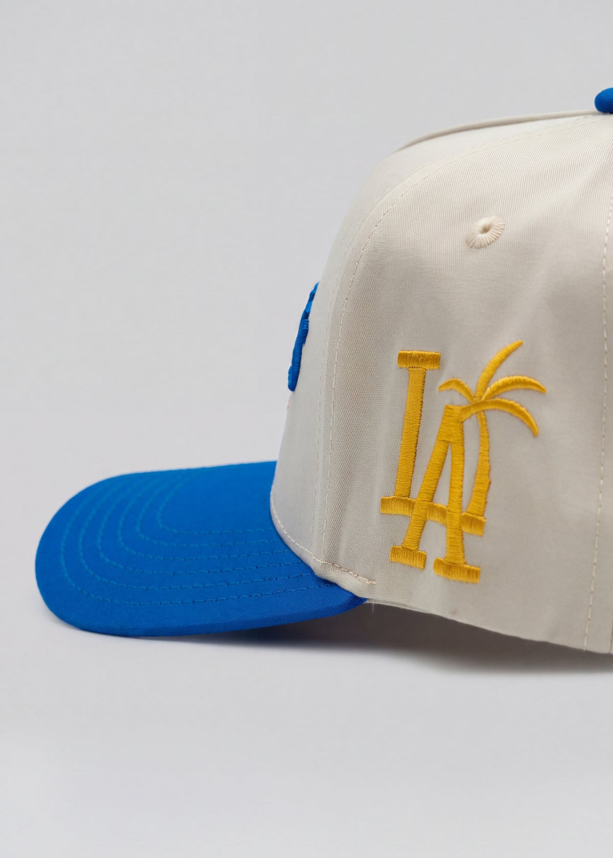 An off-white baseball cap with a bright blue visor and blue side tab, featuring a yellow embroidered 'LA' logo with palm tree details on the front.
