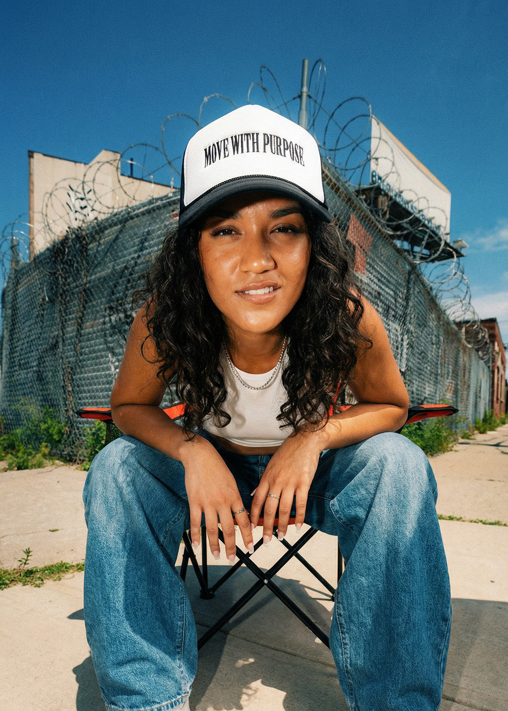 Person wearing a cap with 'Move with Purpose' text, sitting on a chair in an urban setting with barbed wire fence.