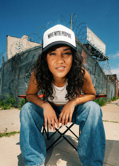 Person wearing a cap with 'Move with Purpose' text, sitting on a chair in an urban setting with barbed wire fence.