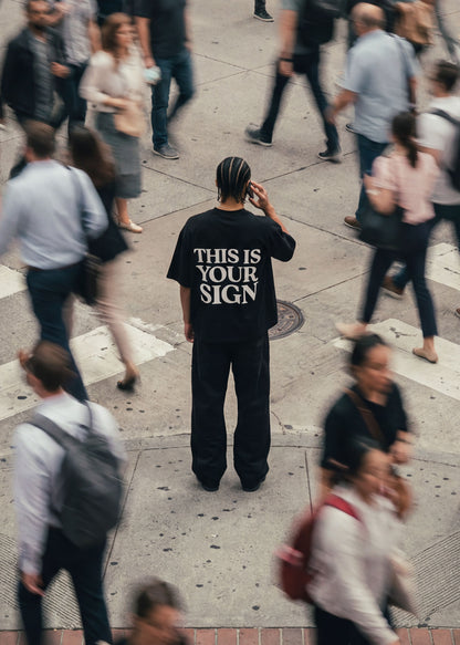 Person wearing a black t-shirt with 'THIS IS YOUR SIGN' on a busy street.
