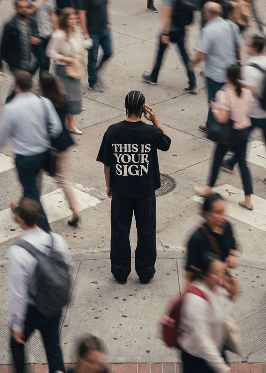 Person wearing a black t-shirt with 'THIS IS YOUR SIGN' on a busy street.
