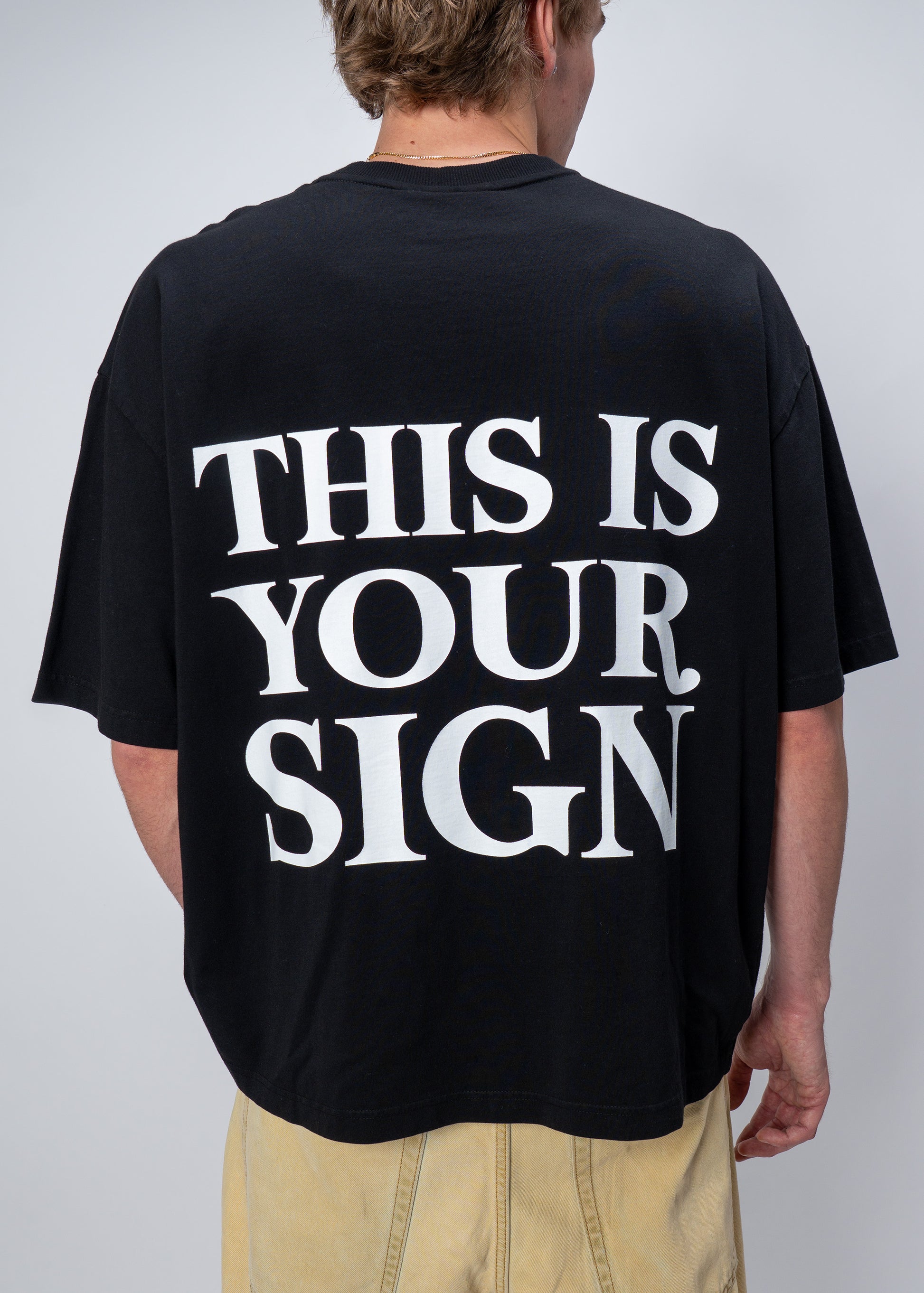 Person wearing a black t-shirt with 'THIS IS YOUR SIGN' text on the back against a plain background
