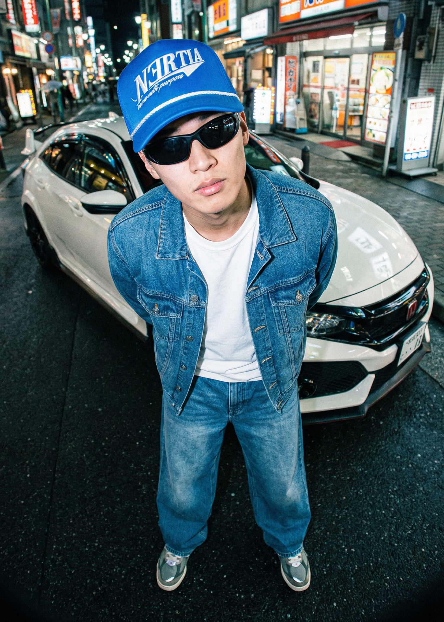 Person wearing a blue cap and sunglasses standing in front of a white car on a city street.