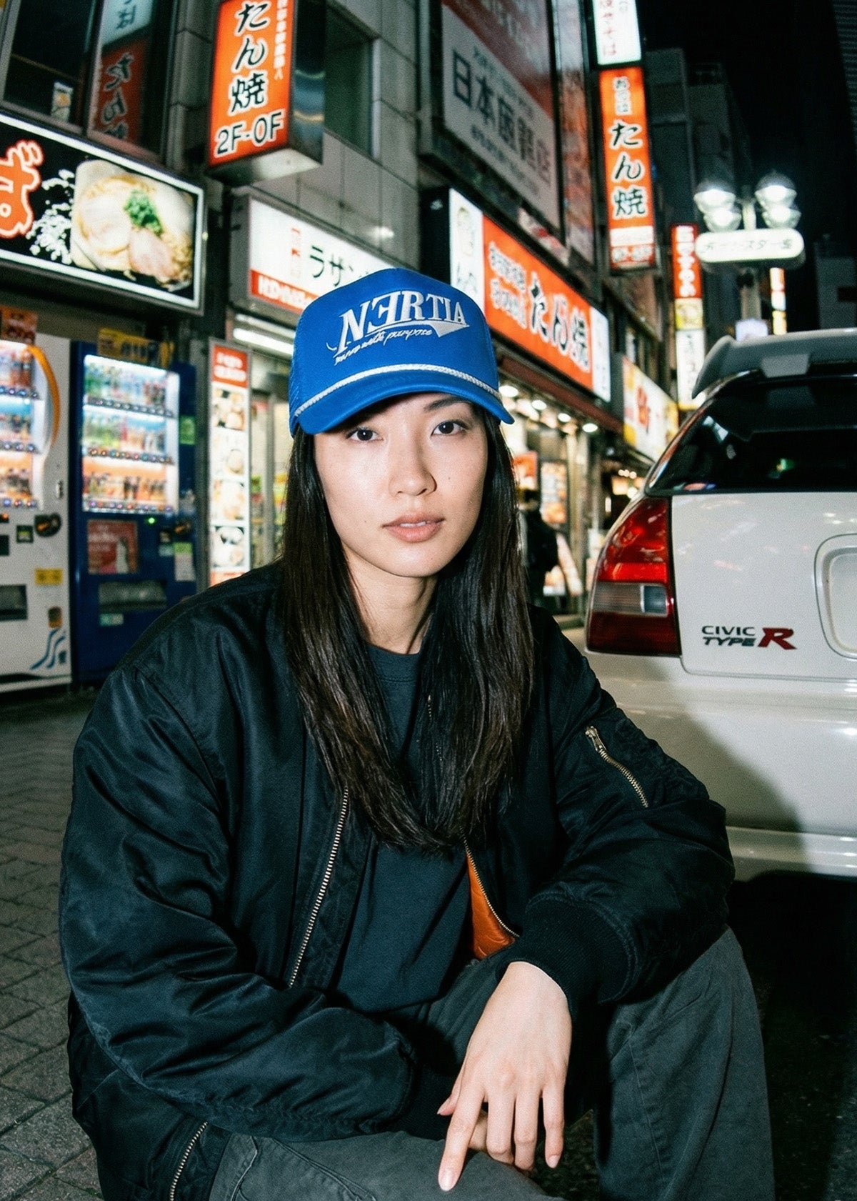 Person wearing a blue cap with 'NCR' logo in an urban night setting with illuminated signs and a car.