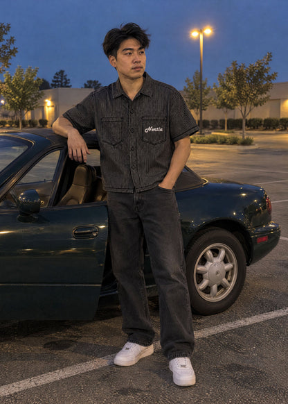 NERTIA Pit Crew Button-Down in black hickory stripe, pocket logo, worn next to a green Mazda Miata in a Torrance parking lot at dusk
